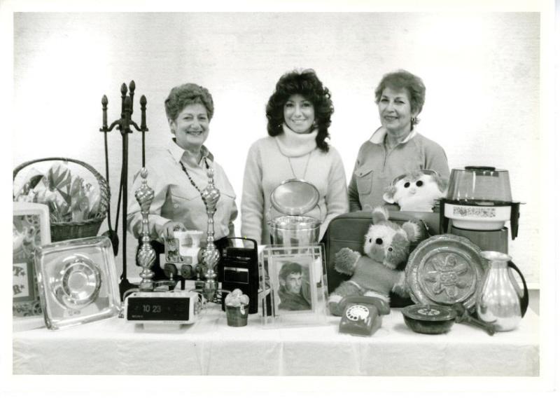  three unidentified individuals standing behind a table displaying a variety of goods, including a clock, fireplace set, photo frame and stuffed animals