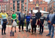 Photo of nine people standing outside in casual clothes. Some are holding red trash pickup tools and white trash bags.
