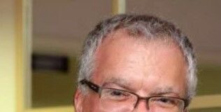 Headshot of an older man with short gray hair and glasses. He is wearing a suit and patterned tie and is smiling at the camera.