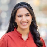 Headshot of a woman with long dark hair smiling at the camera. She is wearing large hoop earrings and a reddish button-down shirt.