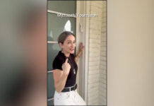 Photo of a woman with her hair pulled back into a ponytail standing outside her front door. She is pointing towards her face and wearing a black T-shirt, beige slacks, a black belt and a silver dog tag around her neck.