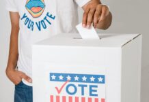 Stock photo of a man putting a folded white piece of paper into a ballot box that says "Vote" in red, white and blue on its front. The man is wearing a white T-shirt that reads "Your Voice, Your Vote" in blue text in a circle around an illustration of an open mouth.