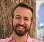 Headshot of a man with short brown hair and a beard and mustache smiling in front of a tree trunk. He is wearing a pink button-down shirt.