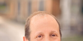 Headshot of a man with short brown hair smiling at the camera. He is wearing a navy blue blazer over a periwinkle button-down shirt. The background is blurred.