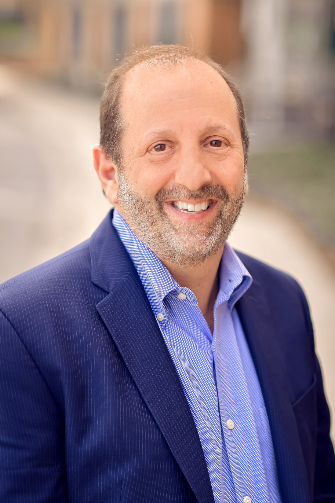 Headshot of a man with short brown hair smiling at the camera. He is wearing a navy blue blazer over a periwinkle button-down shirt. The background is blurred.