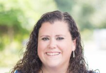 Headshot of a woman with shoulder-length curly brown hair standing outside and smiling at the camera. She is wearing earrings, a long necklace, black eyeliner and a blue T-shirt.