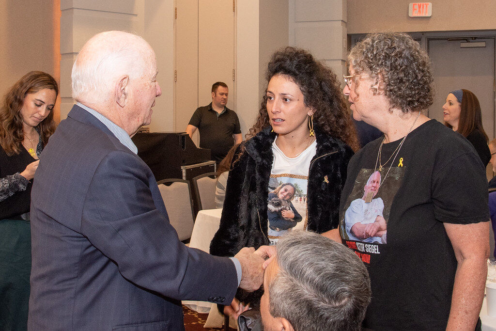 Photo of an older man with gray hair shaking a woman's hand. He is speaking to two women, and the three are looking at one another.