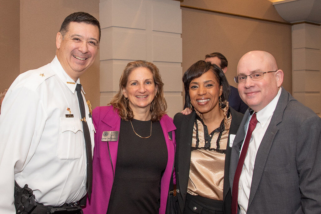 Photo of four adults standing in a row inside: two men and two women, all wearing suits and ties or business casual attire.
