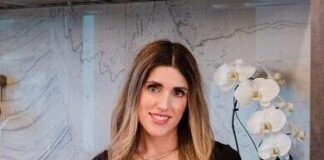 Photo of a woman with long light brown hair, curled on the ends, smiling at the camera. She is leaning on a newly remodeled kitchen counter and smiling at the camera. She is wearing a black shirt and there is a potted white orchid behind her.