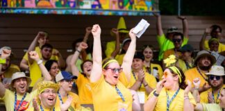 Group photo of dozens of cheering young adults wearing yellow. A woman in the center, dressed in a yellow tutu, has her arms up in the air.