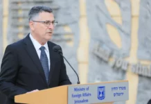 A man wearing glasses, a jacket and a blue tie speaking at a podium.